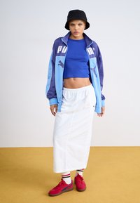 PUMA treningsjakke i blå og lilla, kort blå crop top, hvit skjørt, røde sneakers med blå aksenter, og svart bøttehatt.