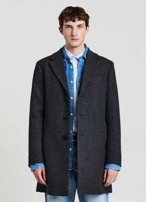 Young man wearing layered dark gray coat over blue denim jacket and light striped shirt, standing against plain light background.