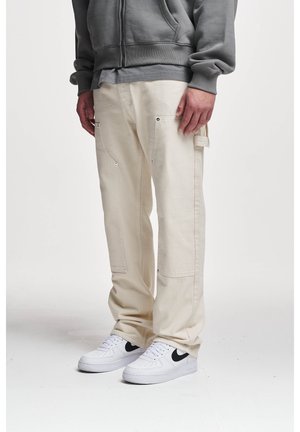 Persoon die crèmekleurige cargobroek, witte sneakers met een zwarte swoosh en een grijze hoodie draagt, staand tegen een effen witte achtergrond.