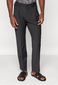 Pantalons plissés gris foncé au fit ajusté, avec une fermeture à bouton et des poches latérales, associés à des sandales à glissière noires.