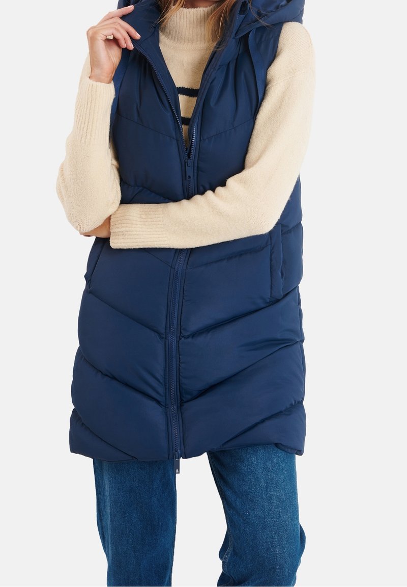 Marineblaue Steppweste mit Kapuze, vorne mit Reißverschluss, gesteppter Optik und Seitentaschen. Wird über einem cremefarbenen Pullover und Jeans getragen.