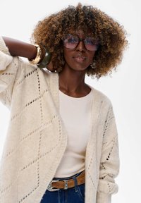 Crème-kleurige gebreide cardigan met diamanten patronen, gelaagd over een beige top en blauwe jeans. Accessoires omvatten grote ronde brillen en armbanden.