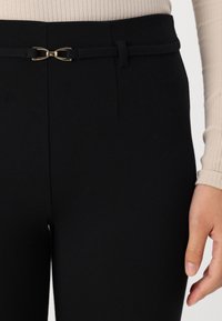 Pantalons noirs taille haute avec une fine ceinture en tissu et une boucle en métal doré. Texture lisse et coupe ajustée.