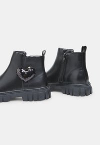 Botas de cuero negras hasta el tobillo con una suela gruesa y un zipper lateral. Presentan un parche en forma de corazón de lentejuelas negras en el lado exterior.