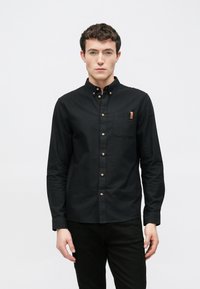 Cămașă neagră cu mâneci lungi și nasturi, cu buzunar pe piept, având nasturi din alamă și un accent din piele pe buzunar. Textură netedă a țesăturii.