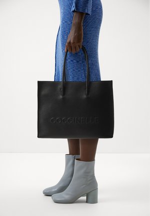 Black leather tote bag with embossed "Coccinelle" logo, featuring vertical handles and a structured shape. Worn with a blue ribbed dress.