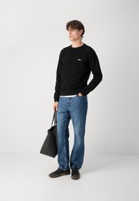 Czarna bluza z białym logo, niebieskie dżinsy i czarne buty. Trzyma czarną torbę na ramię. Minimalistyczny design, luźny krój i casualowy styl.