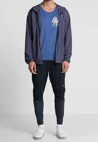 Mężczyzna w granatowej kurtce wiatrówce, niebieskiej koszulce z grafiką, czarnych spodniach sportowych i białych sneakersach stoi z rękami wzdłuż ciała.