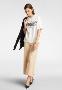 T-shirt bianco in cotone con grafica di farfalla nera, abbinato a pantaloni larghi beige e un cardigan nero, completato da scarpe nere con tacco.