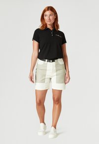 Schwarzes Poloshirt mit weißer Stickerei, kombiniert mit hellbeigen und weißen Shorts. Weiße Sneaker vervollständigen das Outfit.