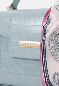 Vaaleansininen krokotiilinattoinen käsilaukku, jossa on kultainen merkkiplakaatti, jossa lukee "ANNA FIELD", sekä kuviollinen silkkihuivi korostuksena sivussa.