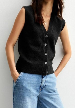 Femme portant un gilet noir sans manches à boutons assorti à un jean bleu clair, les mains posées négligemment dans les poches, sur un fond blanc uni.