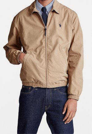 Hombre con una chaqueta beige con cremallera sobre una camisa azul de rayas y corbata azul marino, con vaqueros azul oscuro, posando con una mano en el bolsillo.