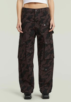 Unterkörper einer Person, die schwarze und dunkelbraune Camouflage-Cargohosen mit mehreren Klappentaschen und schwarze Schuhe mit quadratischer Spitze trägt.