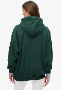 Vrouw met bruin haar, gekleed in een donkergroene hoodie en een witte broek, van achteren gefotografeerd tegen een effen lichte achtergrond.