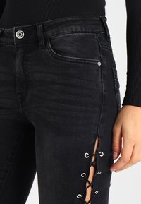 Svarta denimjeans med en elegant design som har en sidoklippning och snörningsdetaljer. Inkluderar två framfickor och dragkedja.