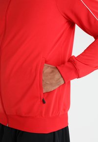 Rode sportjack met een gladde textuur, voorzien van een ritszak en aansluitende manchetten. Eenvoudig ontwerp met contrasterende witte stikselaccenten.