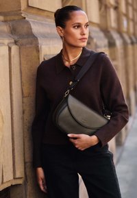 Grå lädercrossbodyväska med en böjd form, guldfärgade detaljer och ett svart justerbart rem, kombinerad med en mörk tröja och svarta byxor.