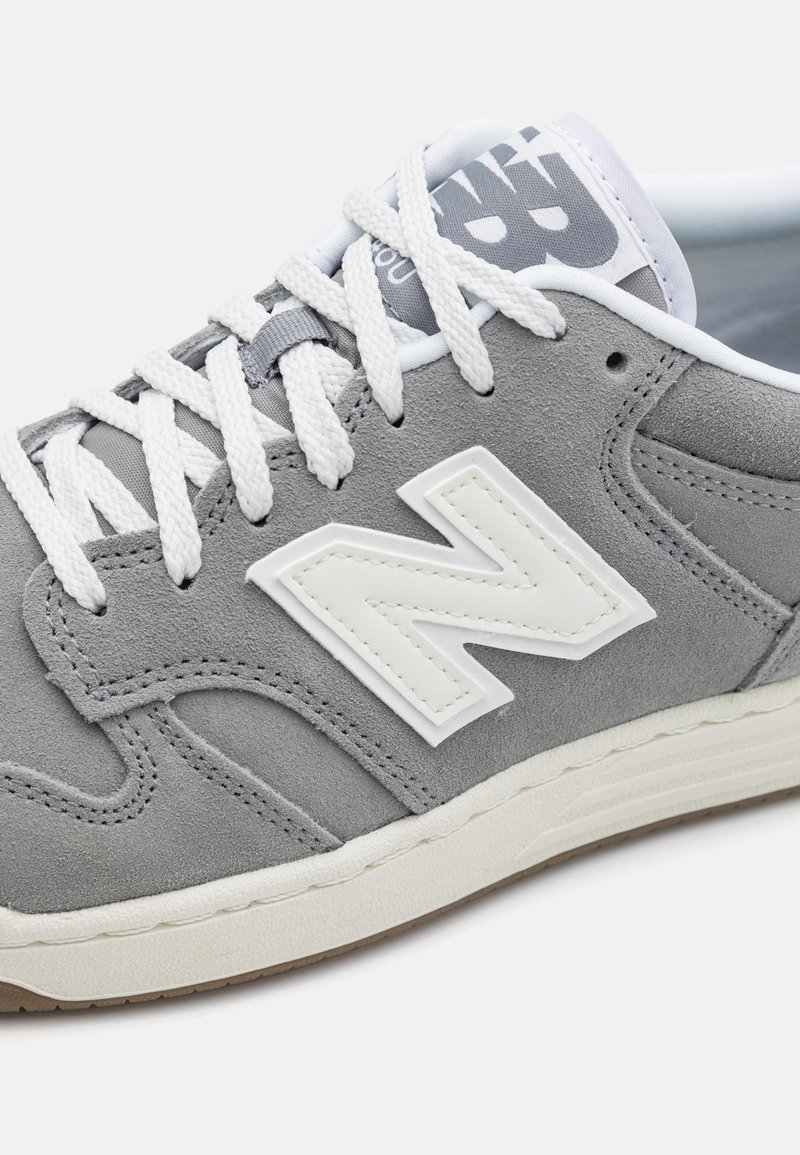 Baskets en suédine grise avec des accents blancs, design à lacets et un logo "N" texturé distinctif sur le côté. Semelle extérieure en caoutchouc visible.