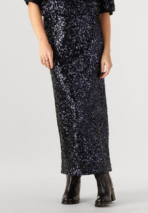 Femme portant une jupe longue noire à sequins associée à des bottines en cuir noires sur un fond clair.