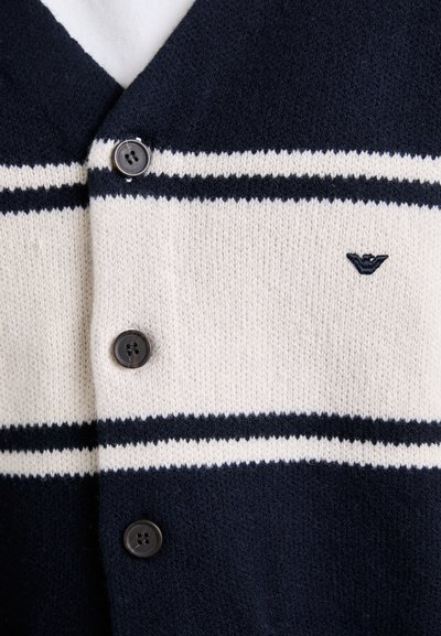 Ναυτικό μπλε και κρέμ ριγέ πλεκτό κασκόλ με τρία σκούρα κουμπιά και ένα μικρό κεντημένο λογότυπο στο στήθος.