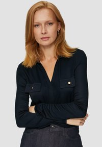 Marineblauwe blouse met lange mouwen en een V-hals, voorzien van twee voorzakken met gouden knopen, gemaakt van een gladde, rekbare stof.