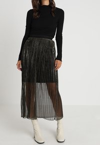 Haut à manches longues noir associé à une jupe plissée en noir transparent avec des accents métalliques. Le mannequin porte des bottines blanches.