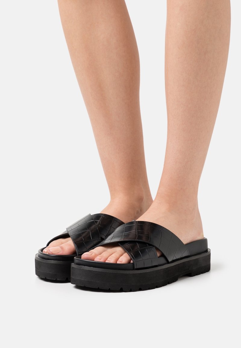 Pieds portant des sandales à plateforme noires avec des brides épaisses croisées à texture crocodile sur fond blanc.