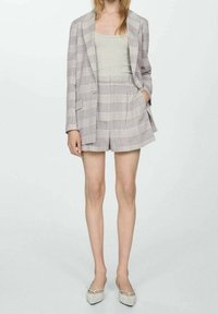 Femme portant un blazer à carreaux gris et un short taille haute assorti, avec un débardeur gris clair rentré à l'intérieur et des chaussures blanches à bout pointu.