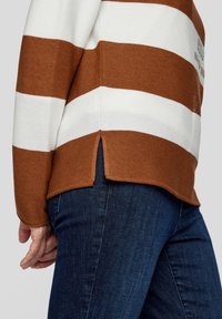 Braun-weißer gestreifter Pullover aus strukturiertem Stoff, mit seitlichen Schlitzen am Saum. Kombiniert mit dunkler Denim-Jeans.