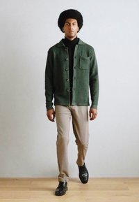 Chaqueta de lana verde con botones y un bolsillo en el pecho, combinada con pantalones beige claros. El modelo lleva zapatos negros y un suéter de cuello alto negro.