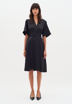 Robe noire avec un design enveloppant, manches larges courtes et une taille ajustée. Texture lisse avec une longueur au genou. Portée avec des talons noirs.