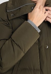 Olivengrøn pufferjakke med quiltet overflade, høj krave, lynlås og trykknaplukninger, lagd under en grå langærmet skjorte.