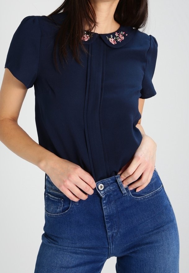 Femme portant un chemisier bleu marine à manches courtes avec broderie florale sur le col, rentrée dans un jean bleu taille haute.