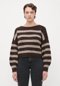 Young person with short curly hair wears a brown and beige striped sweater and dark jeans, standing against a plain white background.