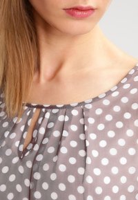 Blusa in tessuto grigio chiaro con pois bianchi, collo tondo e dettaglio a chiave sul davanti, con accenti plissettati.