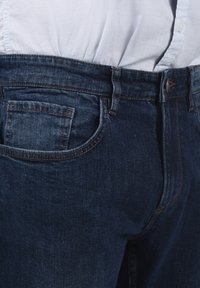 Jeans de mezclilla de color azul oscuro con un corte clásico, que presentan un pequeño bolsillo frontal, costuras discretas y un cierre de botón en la cintura.