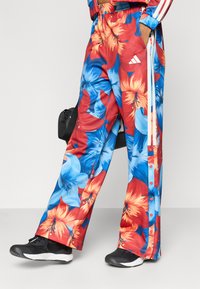 Φαρδιά παντελόνια με λουλουδάτο μοτίβο σε κόκκινο, μπλε και πορτοκαλί, με ελαφρύ ύφασμα. Διαθέτουν κουμπιά και λευκές ρίγες κατά μήκος των πλευρών.