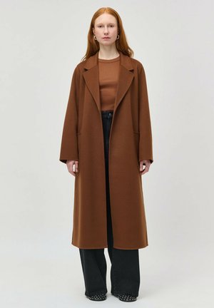 Femeie cu păr roșcat purtând o haină lungă maro, bluză maro, pantaloni largi negri și pantofi negri cu ținte, stând în fața unui fundal simplu.