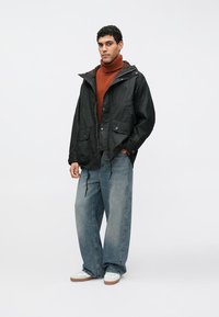 Schwarze wasserdichte Jacke mit Kapuze, zwei Vorderentaschen, über einem orangefarbenen Rollkragenpullover getragen, kombiniert mit locker sitzenden hellblauen Jeans und weißen Turnschuhen.