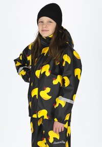 Wasserfeste Jacke mit schwarzem Hintergrund und einem sich wiederholenden Muster aus gelben Gummiente, ergänzt durch reflektierende Akzente und eine Kapuze.