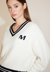Weißer Fleece-Pullover mit V-Ausschnitt, der schwarze und weiße gestreifte Akzente sowie ein gesticktes schwarzes "M" auf der Brust hat.