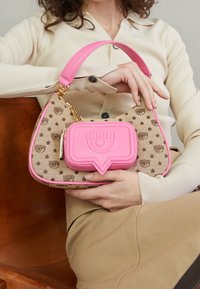 Růžová kabelka se zakřiveným designem, která má tělo z béžové látky zdobené hnědými hvězdami a logy. Výrazný růžový akcent ve tvaru bubliny na konverzaci.