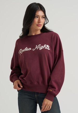 Femme portant un sweat bordeaux avec le texte "Rodeo Nights", associé à un jean foncé, sur un fond gris clair uni.