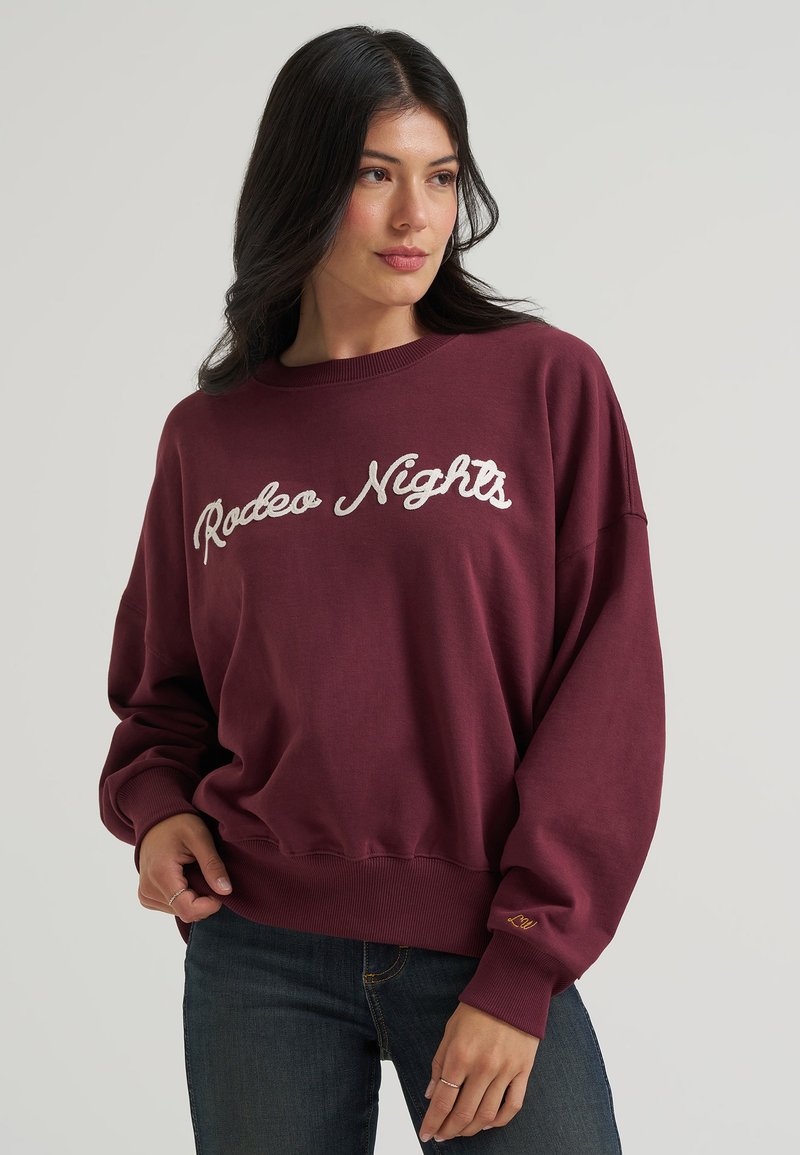 Femme portant un sweat bordeaux avec le texte "Rodeo Nights", associé à un jean foncé, sur un fond gris clair uni.