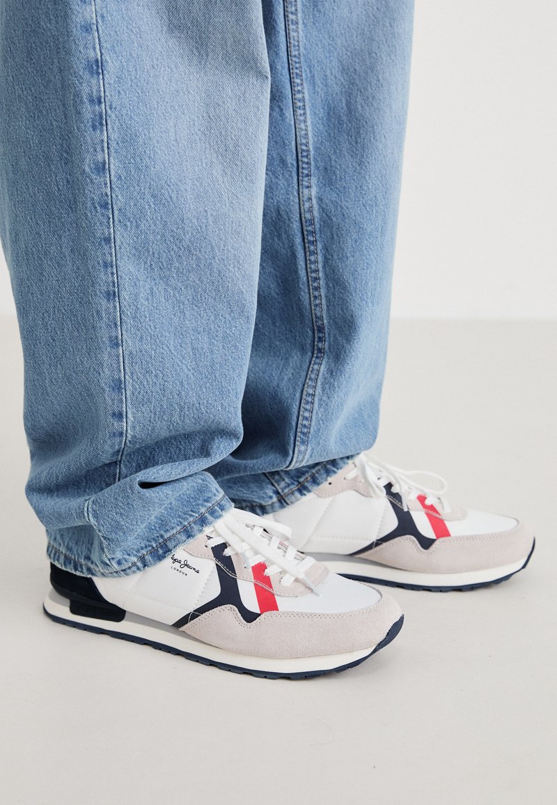 Zapatillas de correr blancas con acentos en gris y azul marino, que presentan una puntera de ante texturizado, cierre con cordones en la parte frontal y un diseño de franjas rojas en contraste, combinadas con jeans azules.