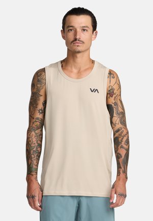 Mann mit Tätowierungen trägt beigefarbenes ärmelloses Hemd und hellblaue Shorts, steht vor einem einfarbigen weißen Hintergrund.