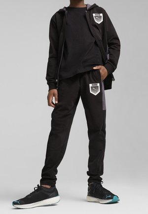 Person iført sort Puma træningsdragt med logopatches på hættetrøje og bukser, kombineret med sort og teal Puma sneakers.