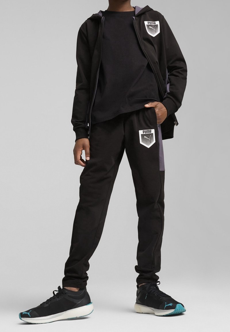 Personne portant un survêtement noir Puma avec des écussons de logo sur le sweat à capuche et le pantalon, associé à des baskets Puma noires et bleu sarcelle.