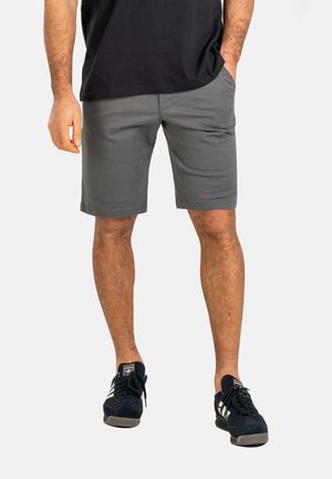 Man wearing gray shorts, black T-shirt, and black Adidas sneakers, standing with one hand in pocket on a plain background.